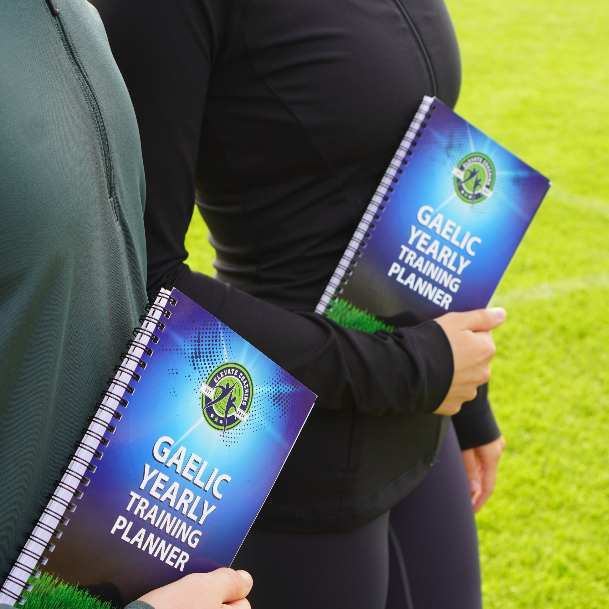 Two people holding Gaelic Yearly Training Planners on a grassy field.